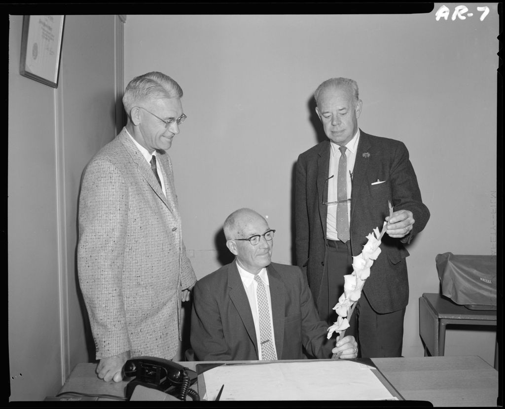 Miniature of Three Men In An Office One Looking At A Gladiola While Other Two Look On